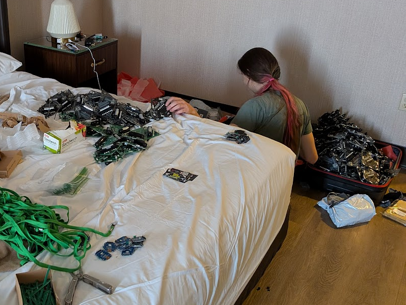 Dani facing away with a giant pile of laser tag badges in a suitcase, and another slightly less giant pile on a hotel bed with various badge parts scattered around