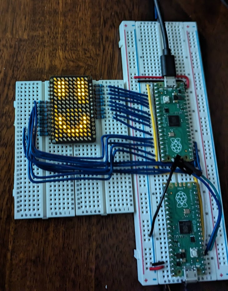 A LED grid on a breadboard with a smiley face pattern illuminated in yellow. It’s connected with 18 blue wires to another breadboard, which has two raspberry pi picos hooked up to it.